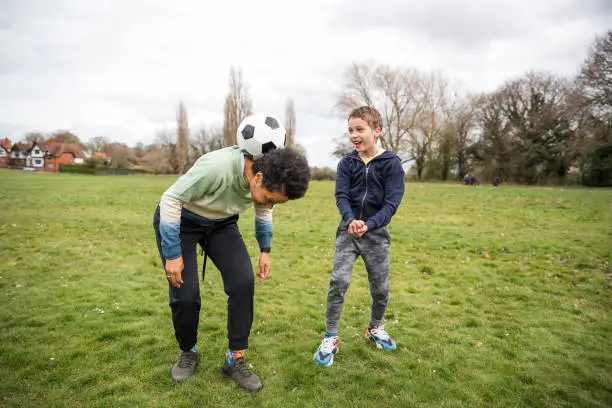 A mature black woman and her son wearing casual sporty clothing in a public park on an overcast day in spring. They having fun and playing with a football.