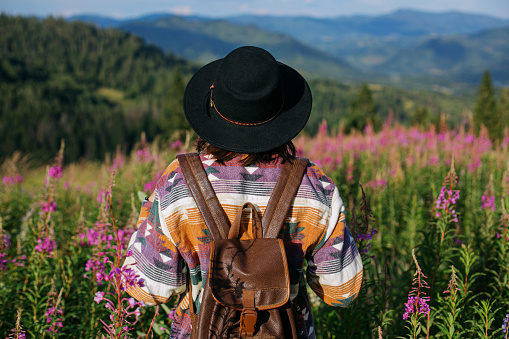 Woman traveler with backpack and hat standing in beautiful wildflowers and looking on mountain hills. Wanderlust and travel concept. Stylish female hiking in mountains, atmospheric moment