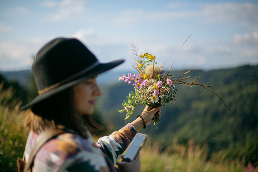 Woman traveler in hat holding bouquet of wildflowers on background of mountain hills and sky. Stylish female hiking in mountains, gathering flowers. Wanderlust and travel concept.