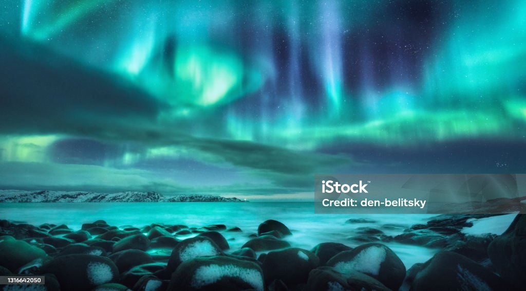 Northern lights. Aurora borealis over ocean in Teriberka, Russia. Starry sky with polar lights and clouds. Night winter landscape with bright aurora, stars, sea, snowy stones in blurred water. Travel Aurora Borealis Stock Photo