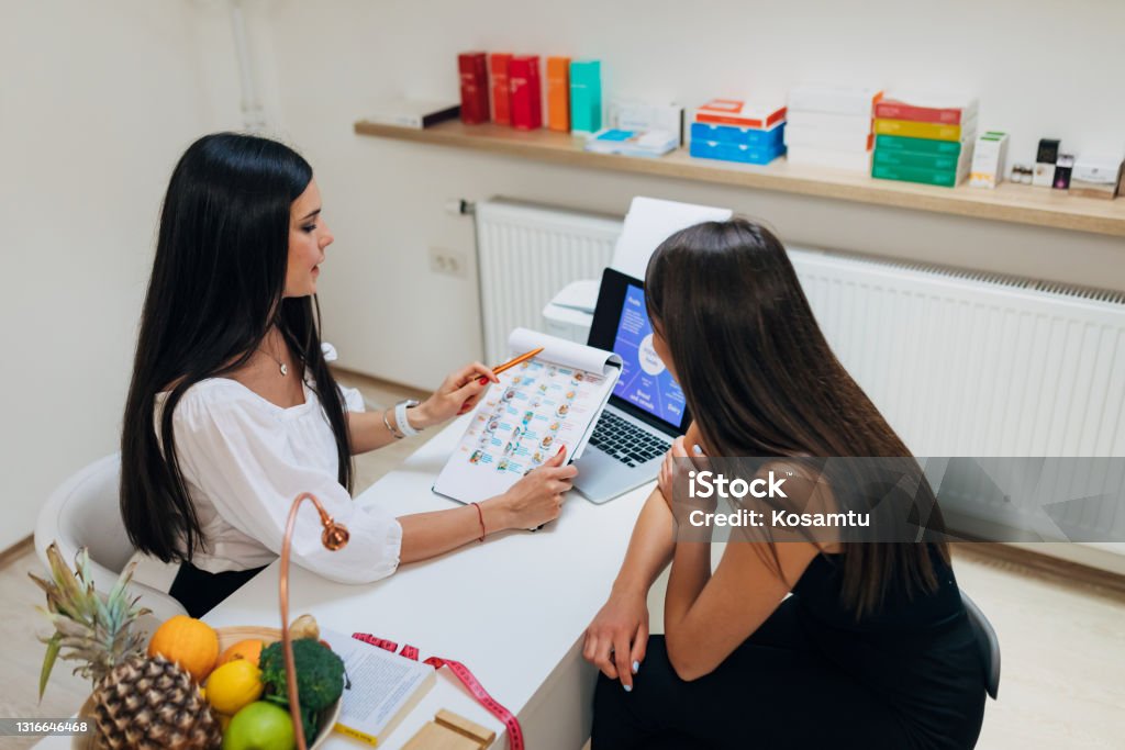 Nutritionist using the chart with food nutrition and calories to explain to her female client how she can control her food portions At the modern doctors office, professional dietologist and nutritionist, showing got her female client the new meal dieting plan, and explaining to her everything she should know Eating Disorder Stock Photo