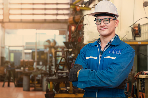Portrait of manual man worker is standing with confident with blue working suite dress and safety helmet in front of indoor factory. Concept of worker operating for production line factory.