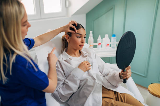 Young woman observing herself in the mirror after the beautician finished procedure of brow lifting with a botox Skilled female plastic surgeon and dermatologist, doing the brow lift procedure on the young female client, while using the botox fillers, in her modern ordination salon mirror stock pictures, royalty-free photos & images