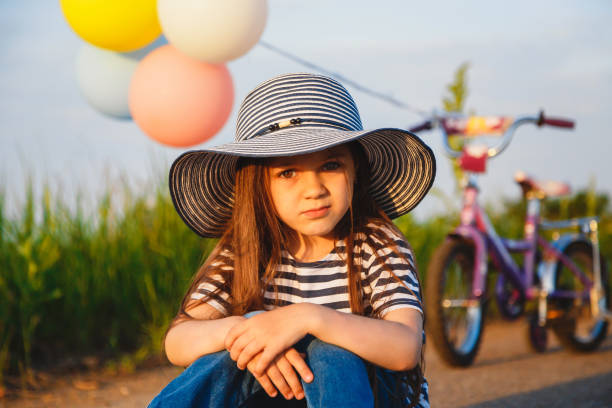 nettes kleines unglückliches mädchen in großen sonnenhut sitzt auf der straße mit ihrem fahrrad hinter ihr - sun sky child balloon stock-fotos und bilder