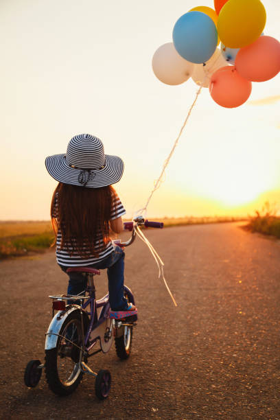 petite fille mignonne avec le vélo coloré de ballons à la route à l’heure d’été de ciel de coucher du soleil. vue arrière - sun sky child balloon photos et images de collection