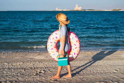 Woman on the beach with a floating doughnut swimming accessory and a hand cooling box for drinks on a sunny day. Summer vacation and seaside fun time abstract
