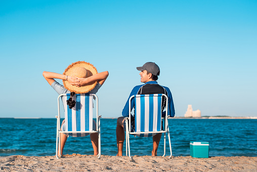 Unrecognizable couple relaxing on the beach while sitting on the tanning chair by the seaside with a small hand fridge cooling box for snacks on a sunny day rear view with copy space. Summer vacation and romantic beach time for two abstract