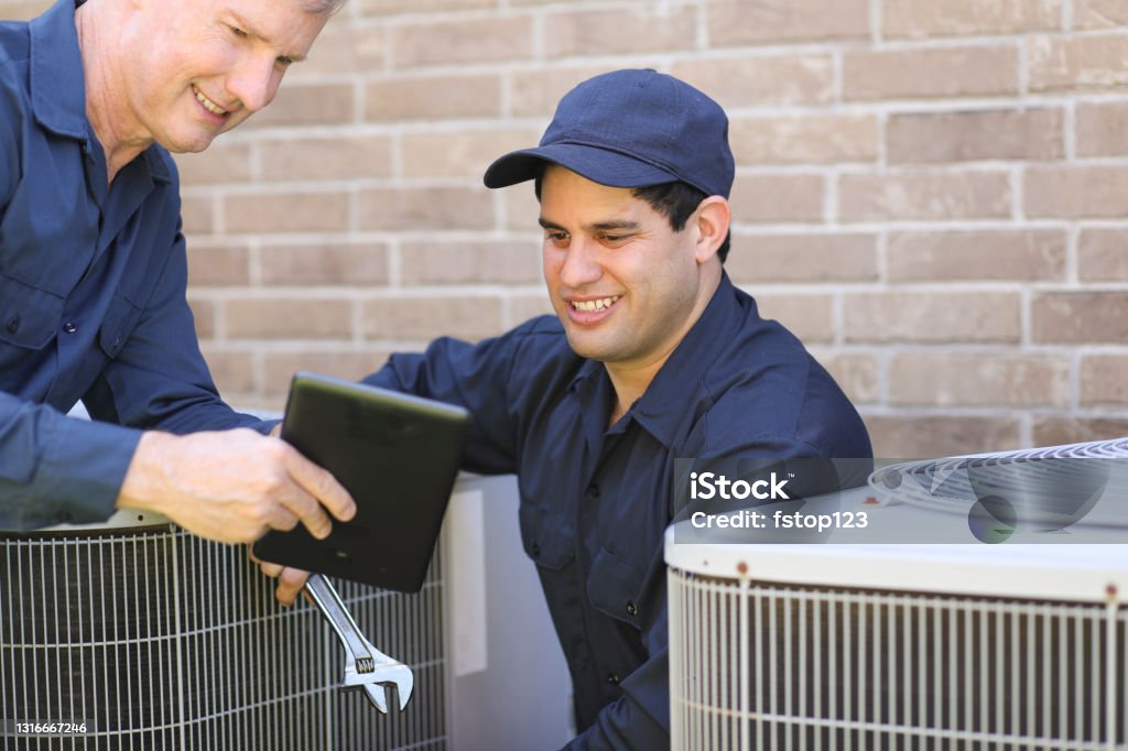 Multi-ethnic team of blue collar air conditioner repairmen at work. Multi-ethnic, mixed age team of blue collar air conditioner repairmen working at residential home.  They prepare to begin work by gathering appropriate tools and referring to digital tablet. Air Conditioner Stock Photo