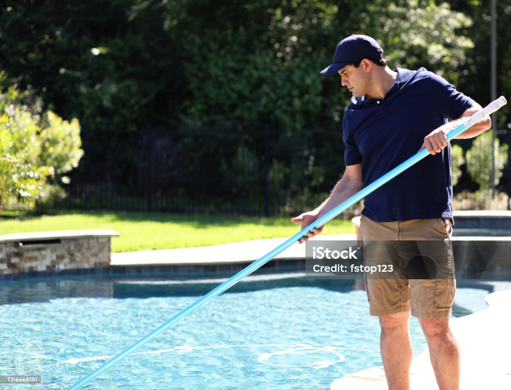 Repairman, cleaning service man at home swimming pool Latin descent man works for local swimming pool cleaning and repair service.  He uses net to clean out pool for summer at home of customer. Swimming Pool Stock Photo