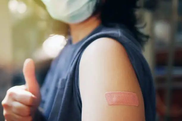 Photo of Close-up adhesive bandage on unrecognized person's arm after injection of vaccine, people in face mask received a coronavirus COVID-19 vaccine and giving thumb up to recommended inoculation vaccine.