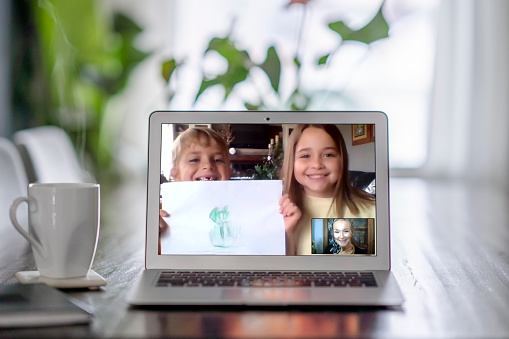A caucasian grandmother video chats with a young boy and girl on a laptop beside a cup of coffee in her home.