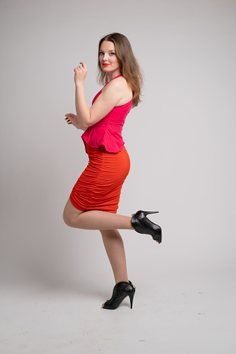 Full length photo of young woman in pink and red dress standing with one leg up against grey background. She is smiling an looking at the camera.