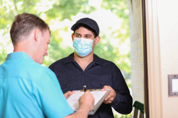 Latin descent repairman, technician makes house call at customer home. Latin descent repairman arrives at customer's front door to make requested home repairs. Mature man greets repairman. He wears a covid mask, blue uniform and holds clipboard and ladder. invoice pad stock pictures, royalty-free photos & images