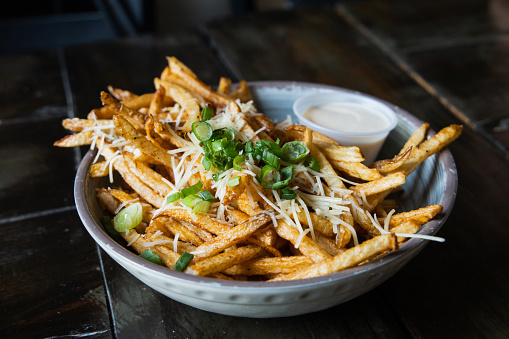 A large bowl of gourmet black truffle french fries in a bowl sprinkled with shredded parmesan cheese, green onions, and a garlic aioli dipping sauce.