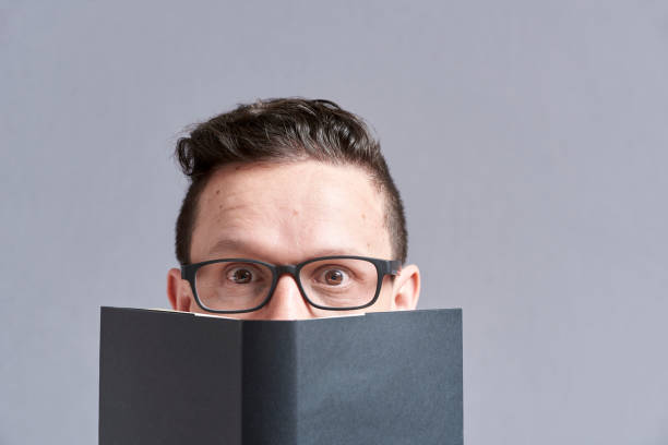 Young man, wearing glasses, looking at the camera, peeking out from behind an open black book. Frontal portrait with gray background and copy space.