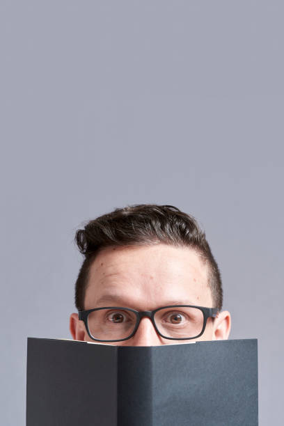 istock Young man, wearing glasses, looking at the camera peeking out from behind a book 1316689160