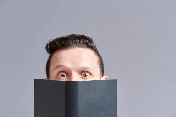 istock Young man staring in surprise at the camera peering out from behind an open book 1316689280