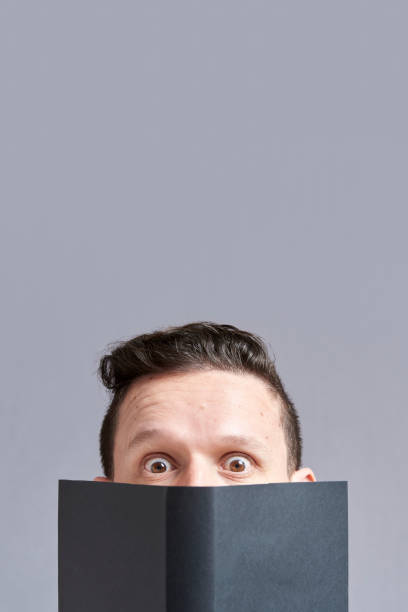 istock Young man staring in surprise at the camera peering out from behind an open book 1316689340