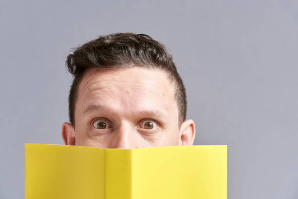 istock Young man looking at the camera, peeking out from behind an open yellow book 1316689390