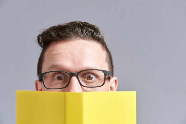 istock Young man, wearing glasses, looking at the camera peeking out from behind a book 1316690055