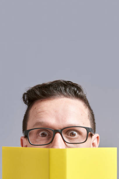 istock Young man, wearing glasses, looking at the camera peeking out from behind a book 1316690114