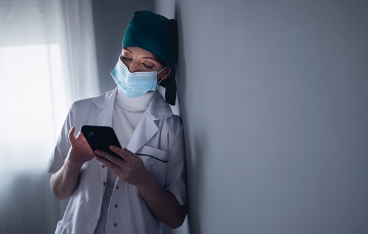 Portrait of tired nurse in medical uniform with surgical cap and surgical mask leaning on the gray wall and using smartphone outside the operating room at medical clinic