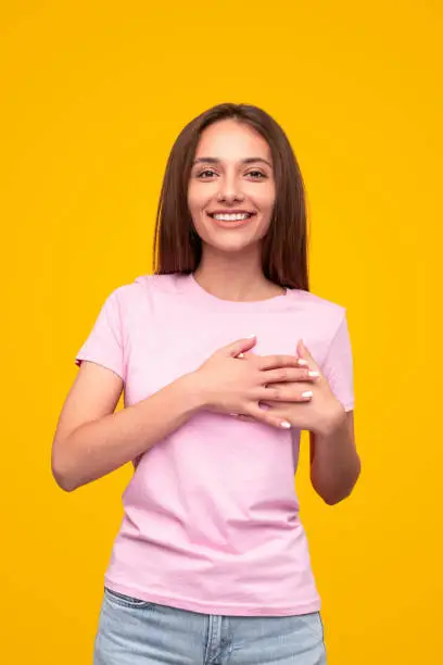 Pleased young woman smiling for camera and keeping hands on chest after hearing compliment against yellow background