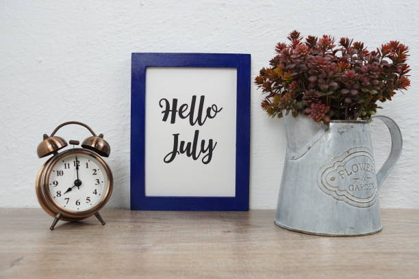istock Hello July typography text with alarm clock and artificial plant decoration on wooden table and white wall background 1316725630