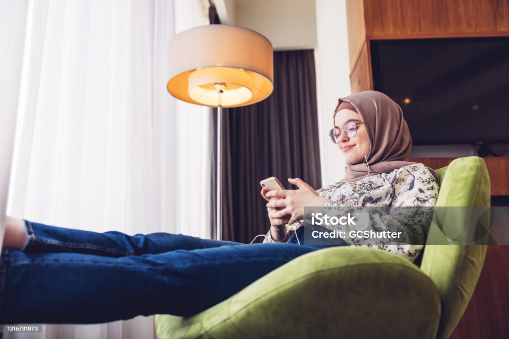 Middle Eastern girl listening to music using smartphone Girl seated comfortably in her hotel room listening to the music using smartphone and earphone Lounge Chair Stock Photo