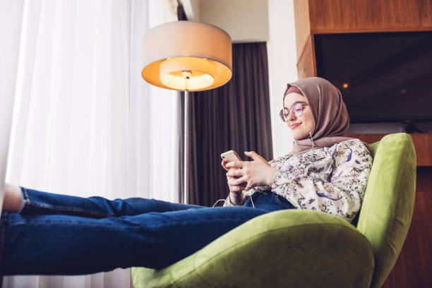 Middle Eastern girl listening to music using smartphone Girl seated comfortably in her hotel room listening to the music using smartphone and earphone chaise longue stock pictures, royalty-free photos & images