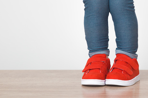 Baby girl legs in blue jeans and red sport shoes on wooden floor at light gray wall background. Closeup. Front view. Empty place for text.