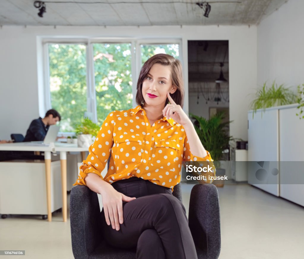 Portrait of woman in the office Mid adult women wearing yellow polka dot shirt sitting on chair in the modern white office, smiling at camera. Yellow Stock Photo