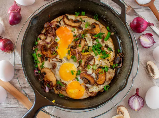 Overhead view of fresh fried eggs with red onions and mushrooms served in a cast iron pan on rustic wooden background