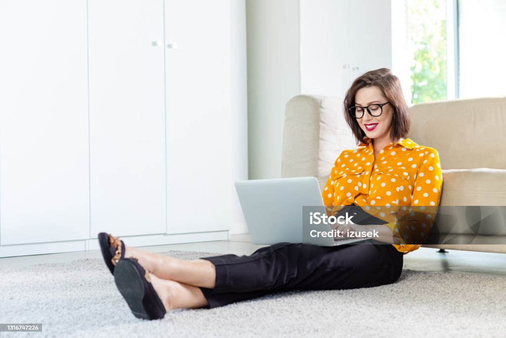 Woman working at home Mid adult women wearing yellow shirt sitting on the floor in her apartment and using laptop. Home office concept. Copywriter Stock Photo