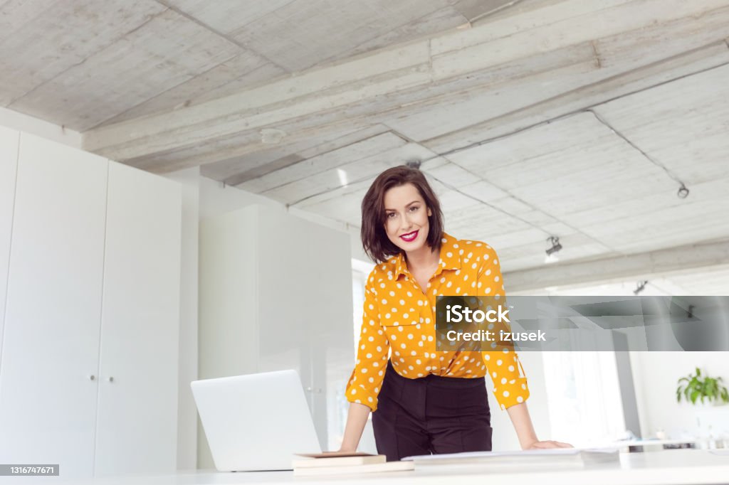 Woman working in the office Mid adult women wearing yellow polka dot shirt standing next to the desk in the modern white office, smiling at camera. Yellow Stock Photo