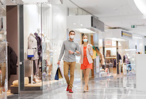 couple in medical masks with shopping bags in mall sale, consumerism and pandemic concept - happy young couple wearing face protective medical mask for protection from virus disease with shopping bags walking in mall mall walker stock pictures, royalty-free photos & images