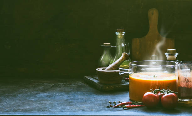 요리 재료와 어두운 벽 배경에 소박한 테이블에 유리 냄비에 증기와 뜨거운 토마토 수프. 부엌 장면, 정물. - tomato soup red basil table 뉴스 사진 이미지