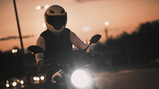 Portrait of an young man sitting on motorcycle and wearing a leather jacket and helmet. Night time. Low light. Traffic jam.