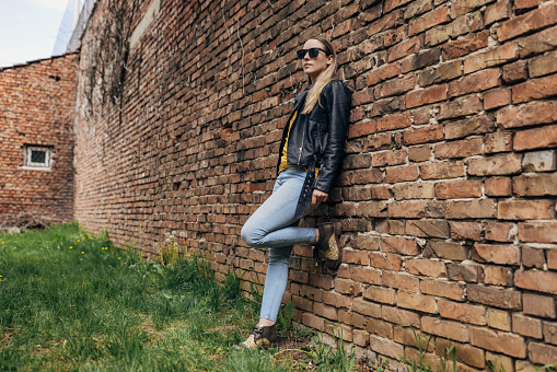 One woman, female in modern clothing standing by a brick wall outdoors.