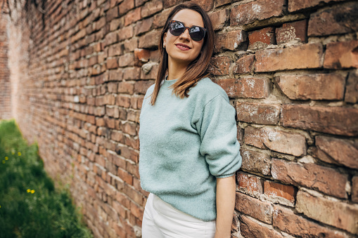 One woman, female in modern clothing standing by a brick wall outdoors.