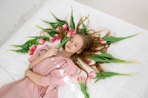 A beautiful long-haired girl is lying in bed, surrounded by pink tulips.