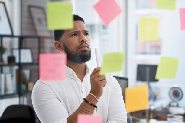 scatto di un giovane uomo d'affari che si divaga con note appiccicoso su una parete di vetro in un ufficio - decisions foto e immagini stock