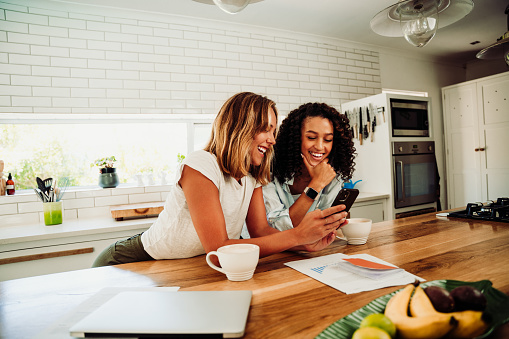 Mixed race female students taking work break chatting in kitchen with cellular device and hot coffee bonding together. High quality photo