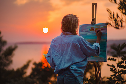 Rear view of a Caucasian female painter painting her art with an impressive and beautiful landscape of a coastline with a sunset in the background using an easel set up on a porch