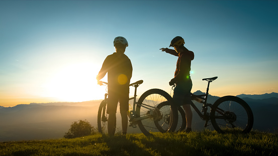 Two silhouettes standing near mountain bikes, talking and observing view in the background, mountaintops