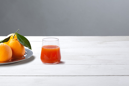 Orange Juice Drink in Glass on White Wooden Table and Gray Background – Whole Oranges on Silver Plate, Green Leaves, with Shadows, Red Juice – High Quality Minimalist Ingredient Photo