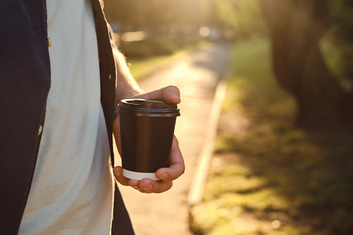 Black paper cup with coffee in man hand. Time for drinking coffee in the city. Disposable paper cup closeup. Blank space for text, mockup. Concept of coffee to go, takeaway drinks, morning snack