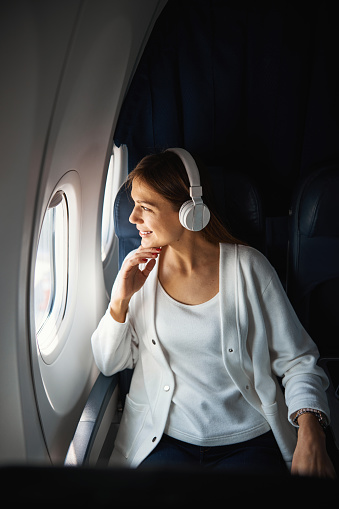 Mirthful pretty woman smiling while sitting by the airplane porthole with headphones on her head