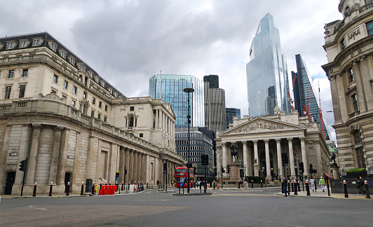 Bank of England (left) off Threadneedle Street with red London double decker bus (ahead) and Royal Exchange (right) background of city skyscrapers. Outdoors on an overcast summers day.  May 4, 2021