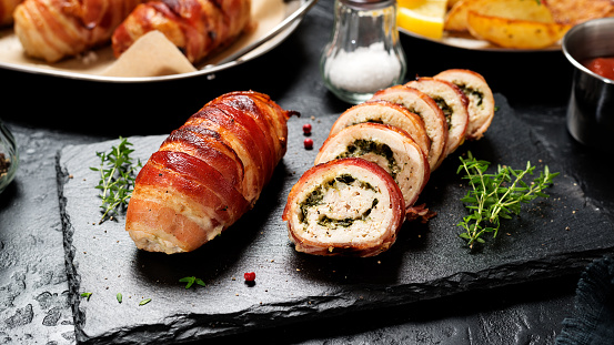 Bacon wrapped chicken rolls stuffed with feta cheese and spinach on black stone background. Beautiful festive lunch or dinner.